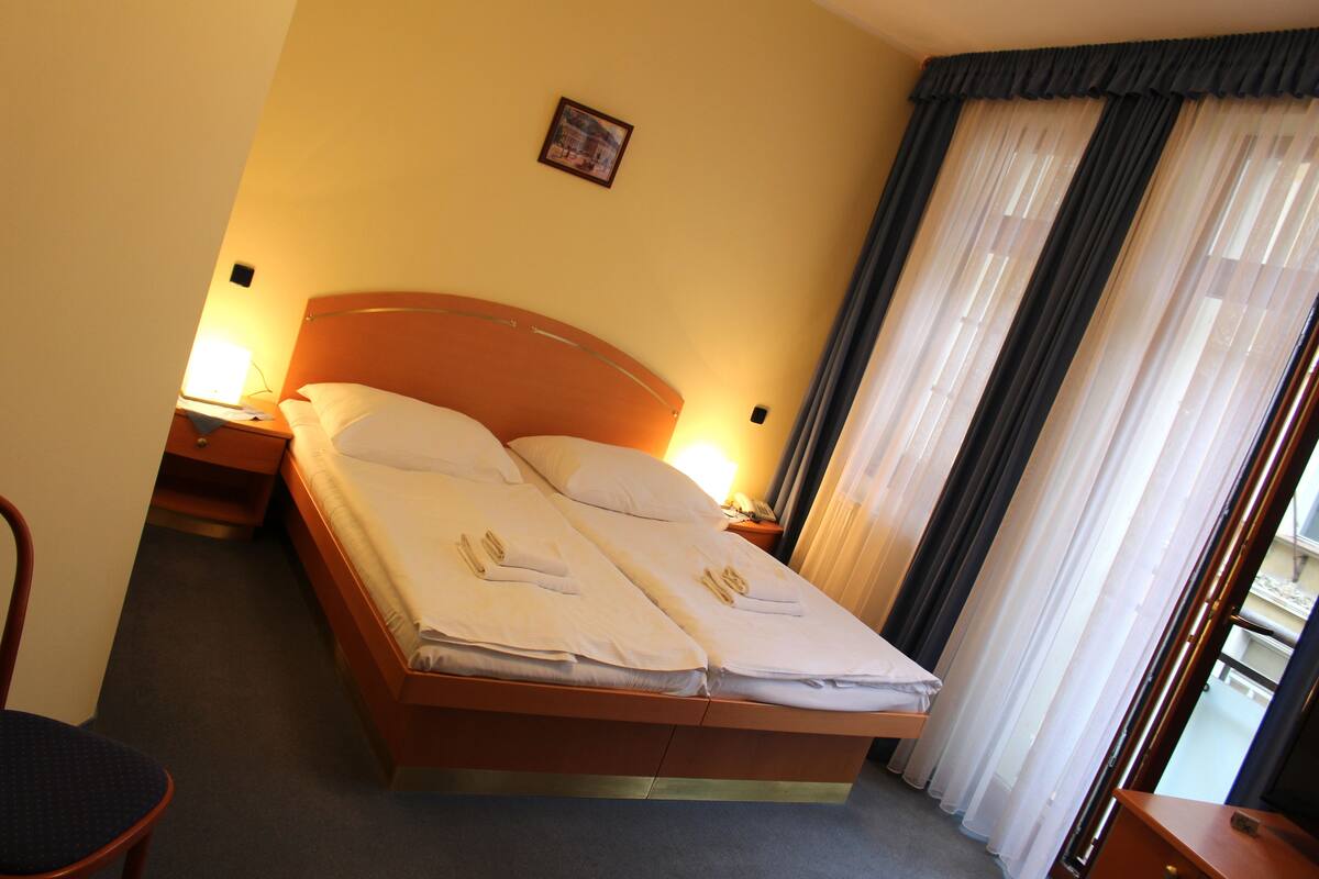 A double bed with white linens, two bedside tables with lamps, a chair, and a window with sheer curtains.