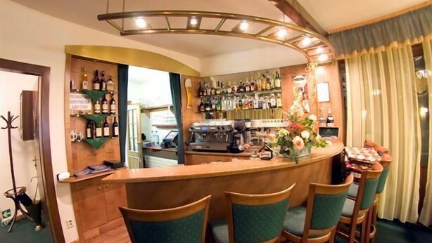 A bar area with a curved counter, wooden chairs, and a view of the kitchen in the background. - Hotel Ruze (Karlovy Vary)