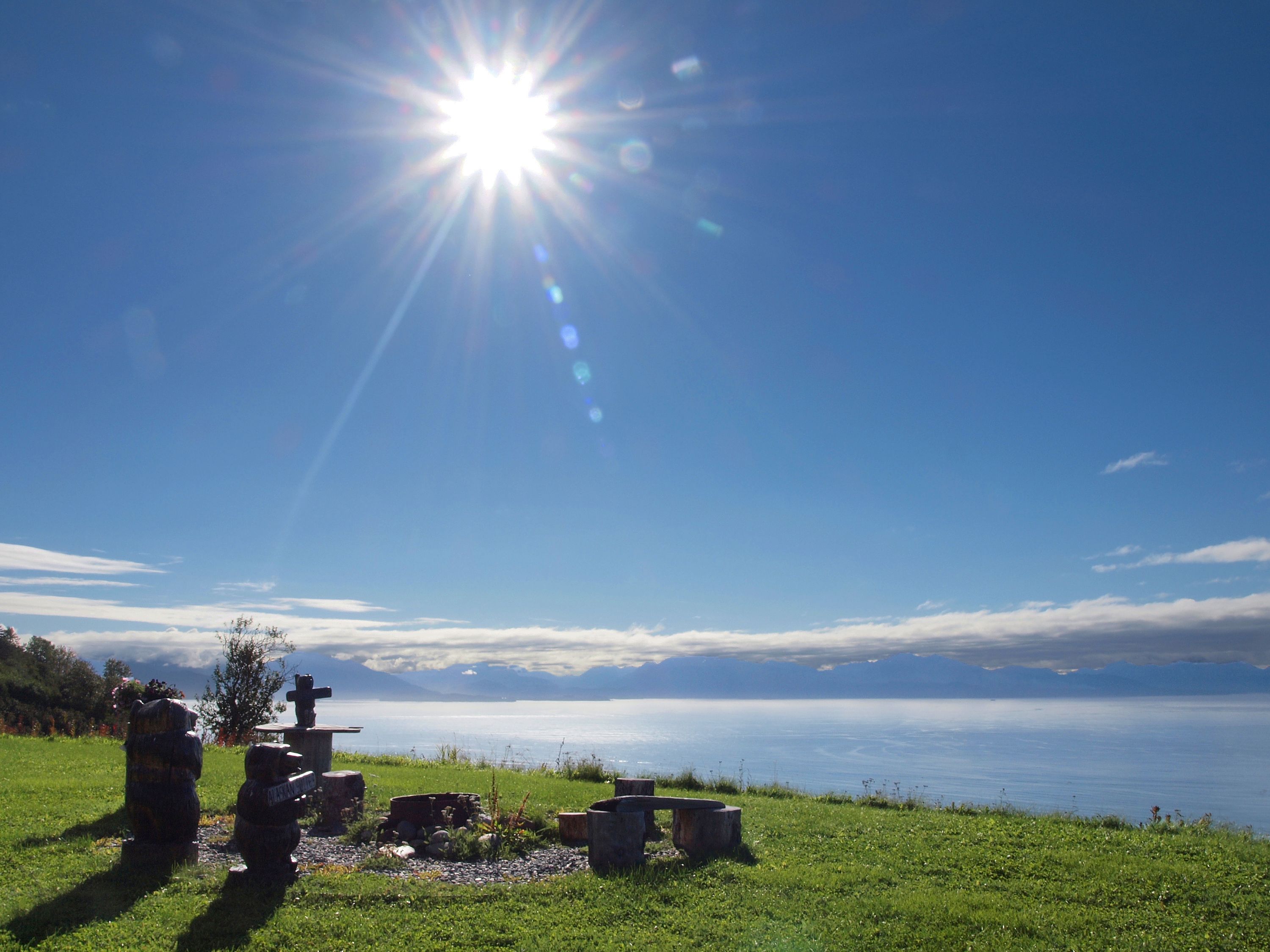 A grassy area with a stone bench and a cross, overlooking a body of water and distant mountains.