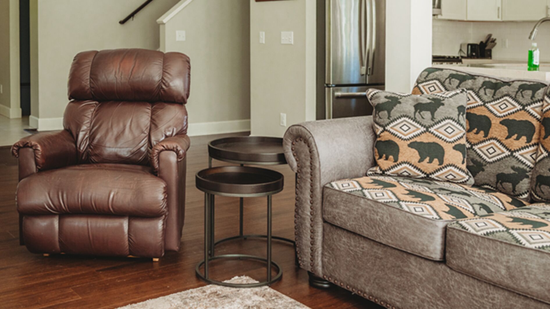 A living room with a brown leather recliner, a gray sofa with patterned cushions, and two small side tables.
