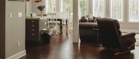 A modern living room with a brown leather chair, a dining area with white chairs, and a kitchen with a white countertop and a trash can.