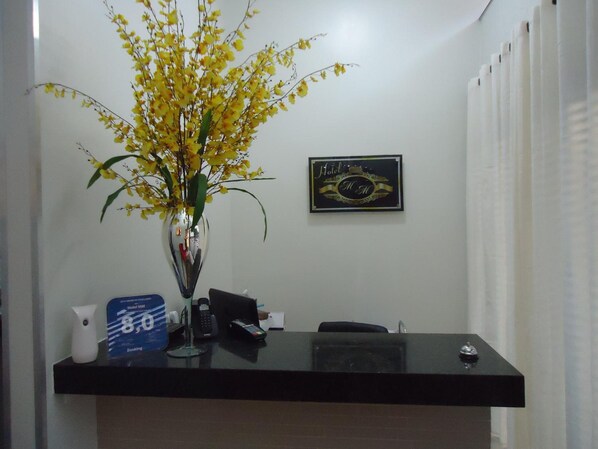 Un mostrador de recepción de hotel con un jarrón de flores amarillas, un cartel enmarcado y un atril azul para reservas.