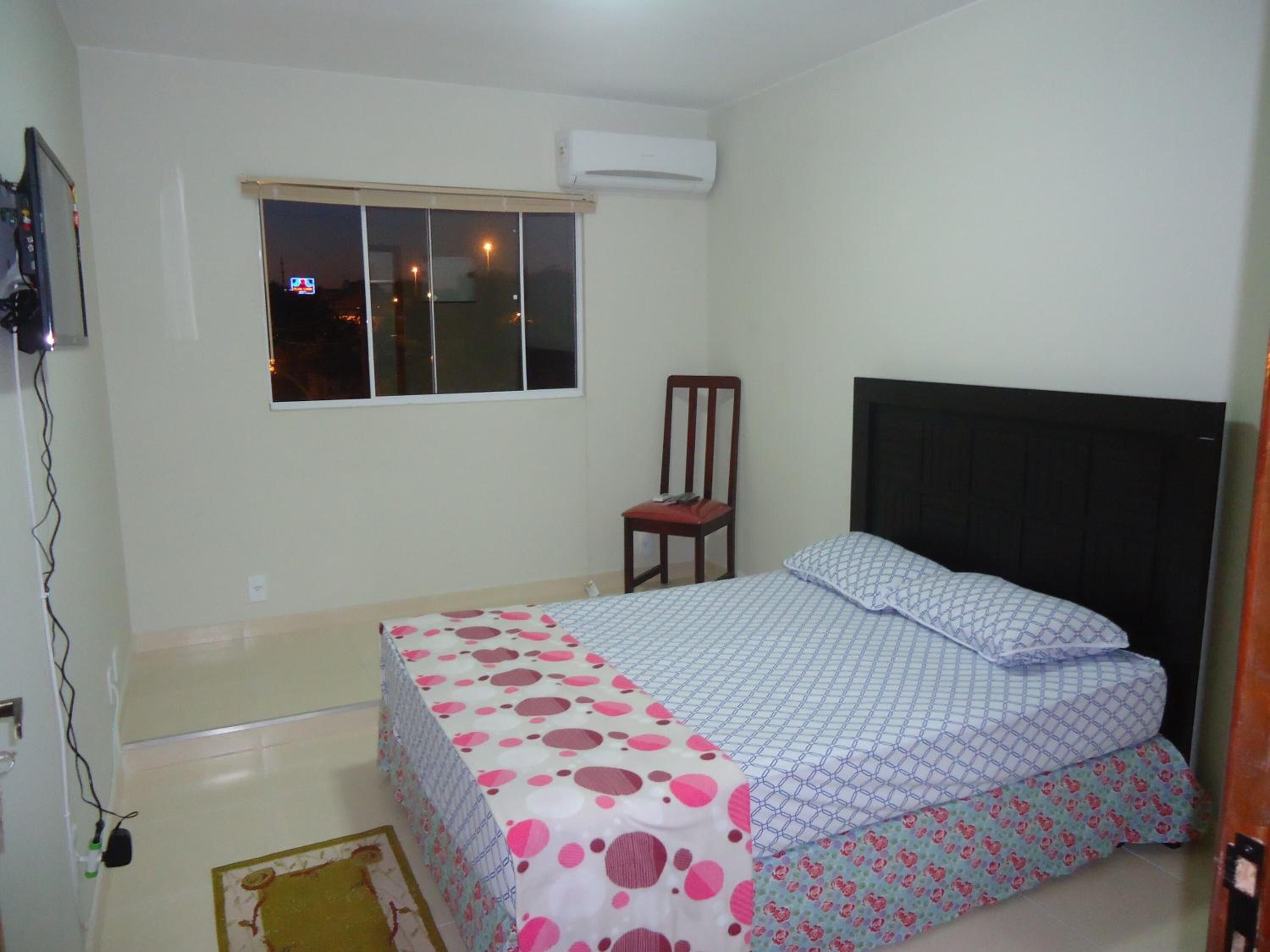 A bedroom with a bed, a chair, a window, and an air conditioning unit.