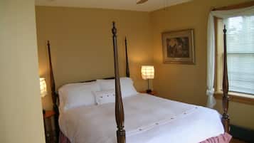 A four-poster bed with white linens, two bedside lamps, and a ceiling fan in a room with beige walls and a window with blinds.