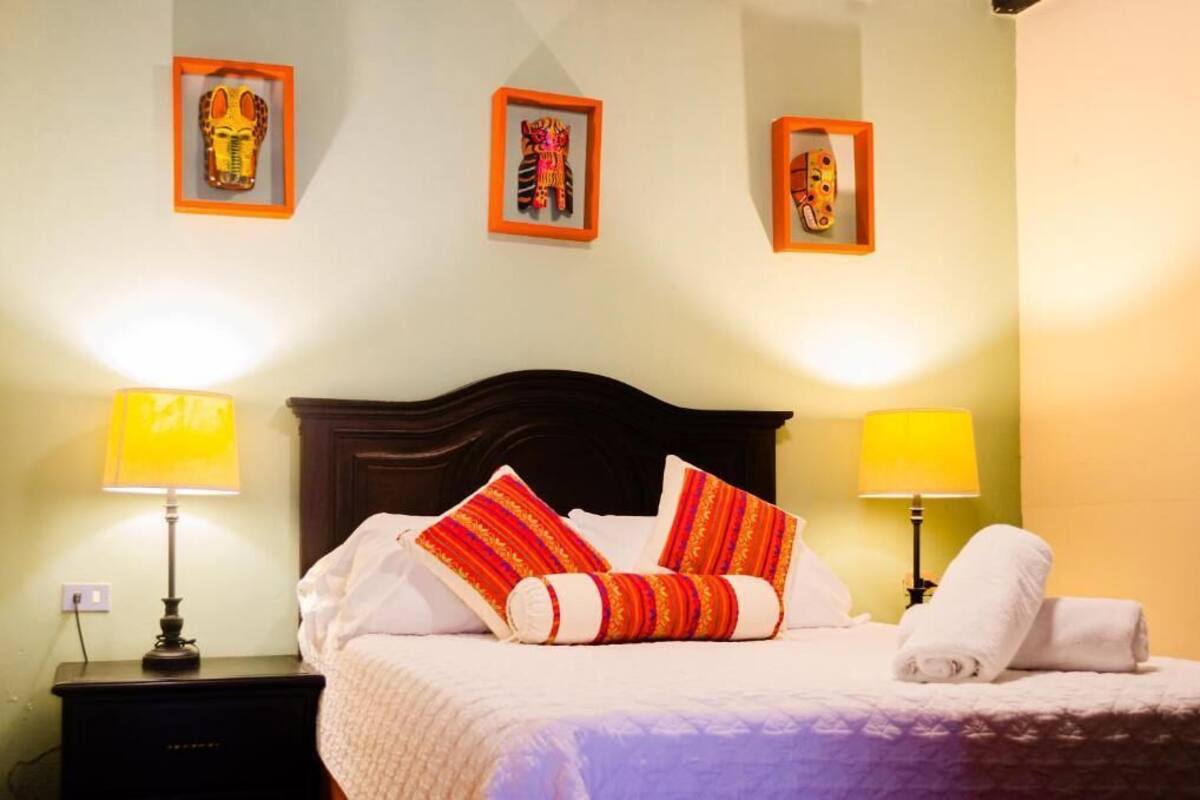Una cama bien hecha con almohadones a rayas, dos lámparas de noche y cuadros en la pared.