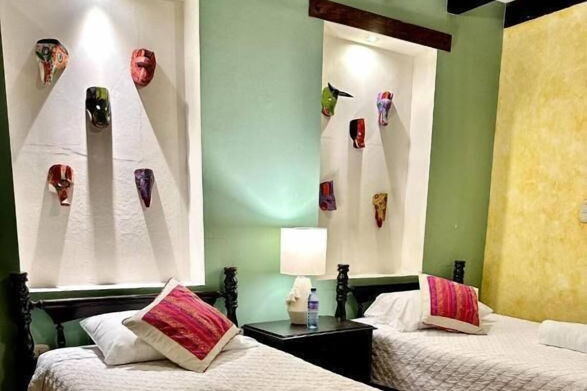 Habitación de hotel con dos camas, cada una con colcha blanca acolchada y una toalla enrollada. Los cabeceros están sujetos a la pared y decorados con máscaras.