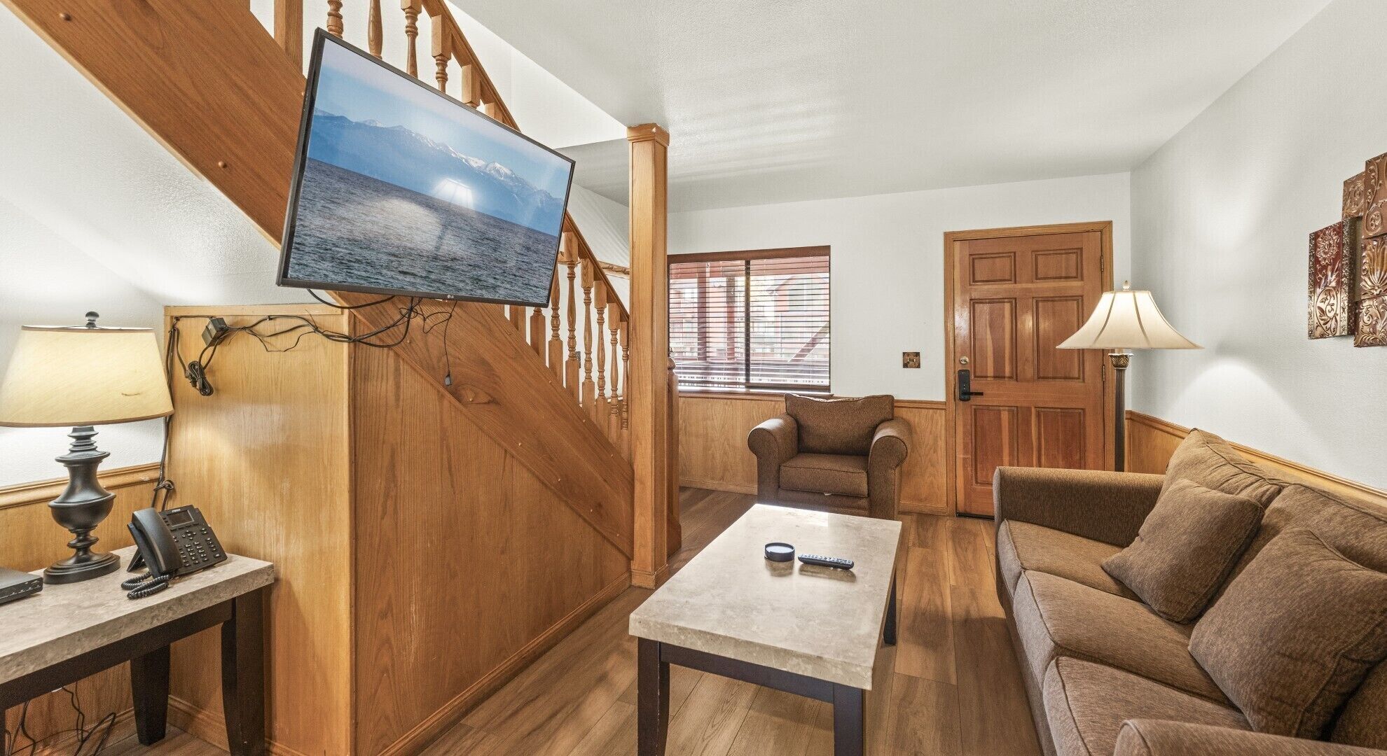 Una sala de estar con una escalera de madera, un televisor montado en la pared, un sofá, una mesa de centro y una lámpara.