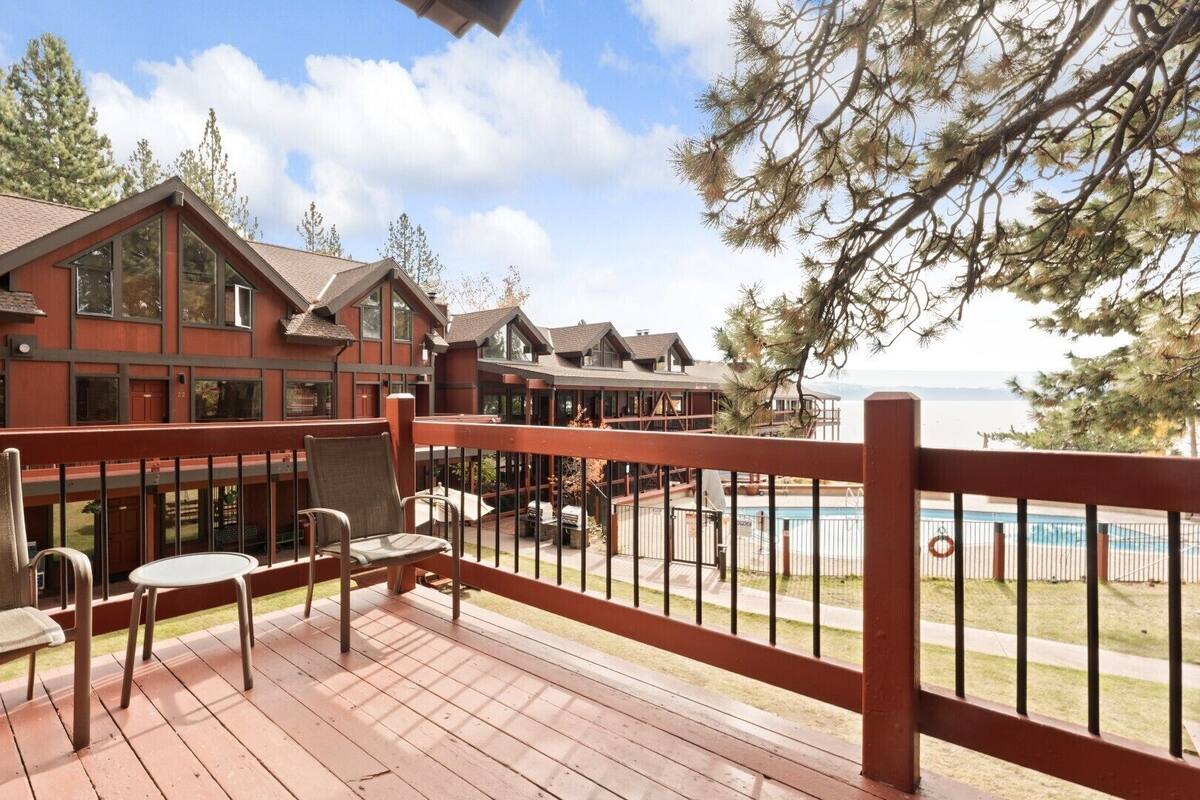A wooden deck with chairs and a table, overlooking a pool and a building with multiple windows.