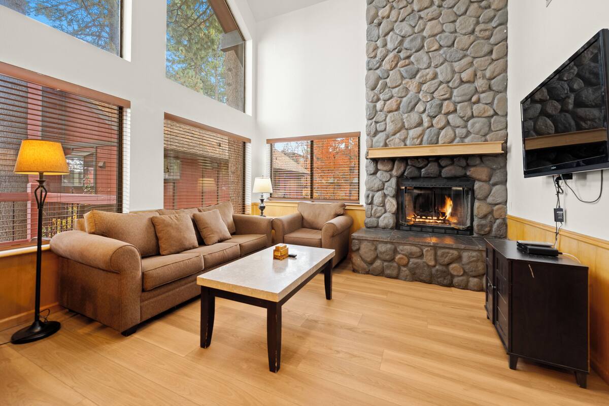 A cozy living room with a stone fireplace, a sofa set, a coffee table, and a TV mounted on the wall.