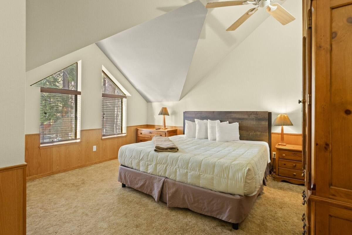 A bedroom with a sloped ceiling, a large bed, two bedside tables with lamps, and windows with blinds.