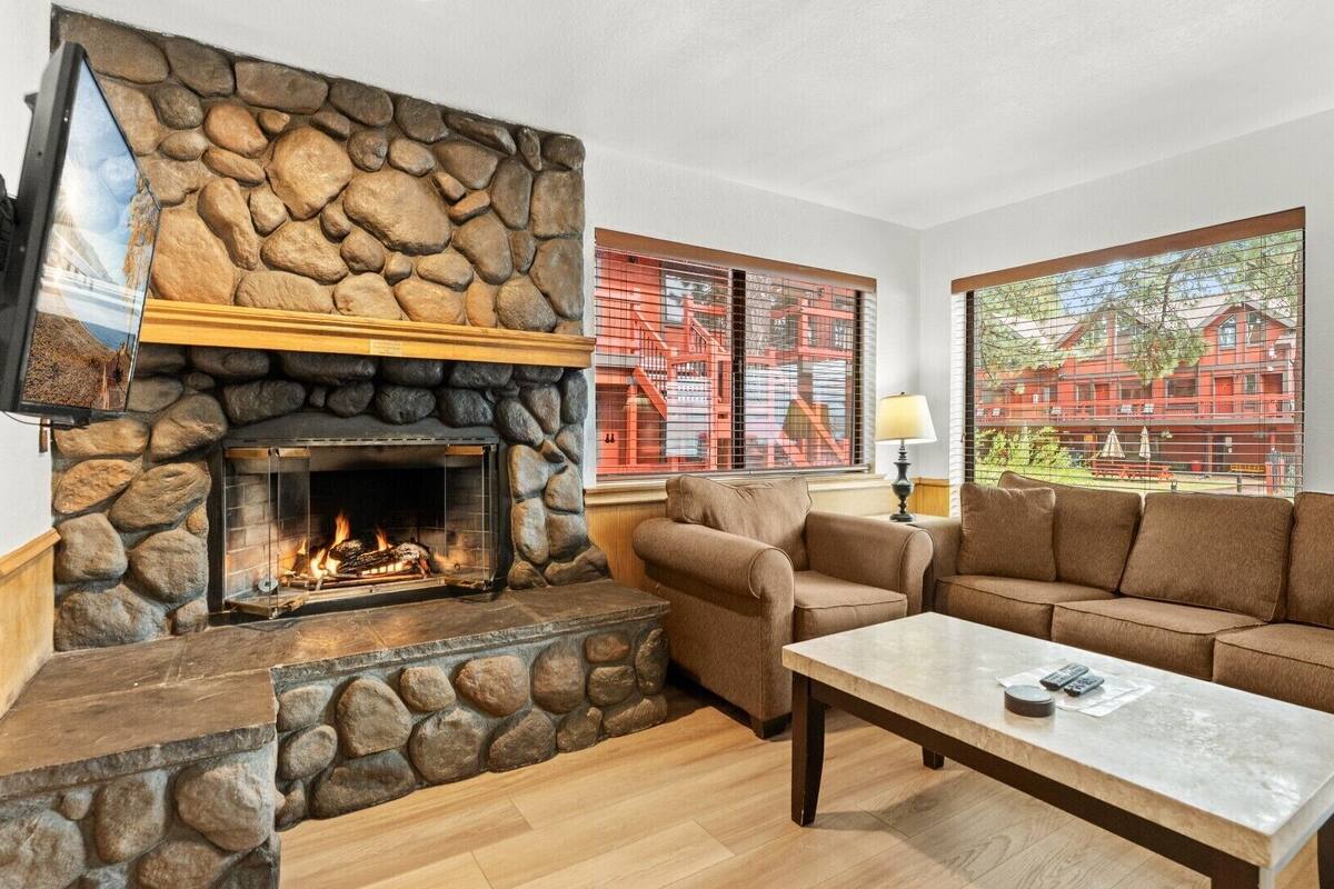 A living room with a stone fireplace, a sofa, a coffee table, and large windows with a view of red buildings.