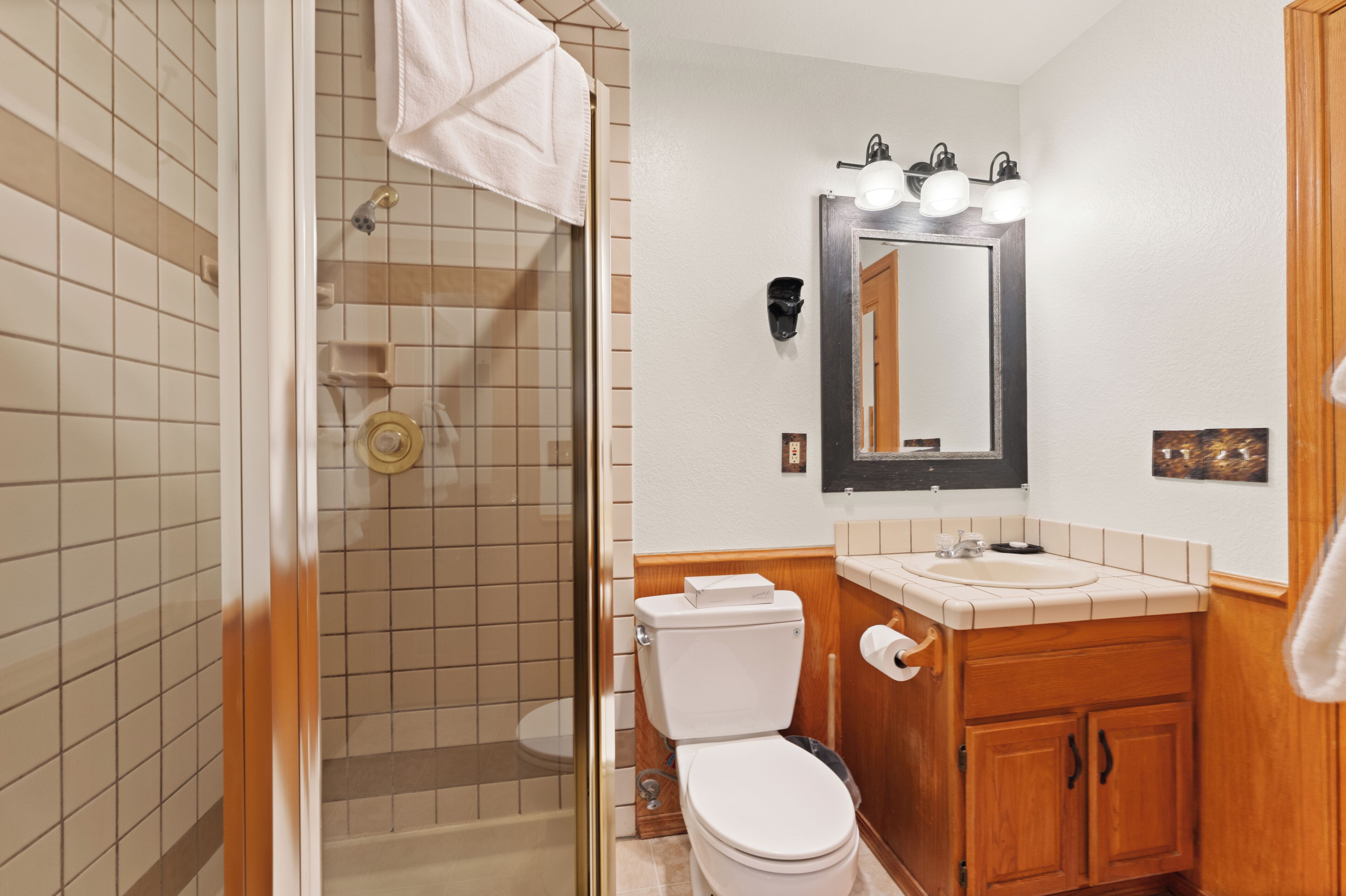 A bathroom with a glass-enclosed shower, a white toilet, a wooden vanity with a sink, and a mirror with lights.