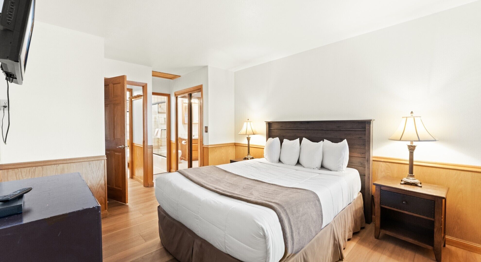A hotel room with a large bed, two bedside tables with lamps, a desk with a remote control, and a TV mounted on the wall.