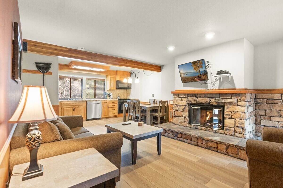 A spacious living area with a stone fireplace, wooden furniture, and a kitchen in the background.