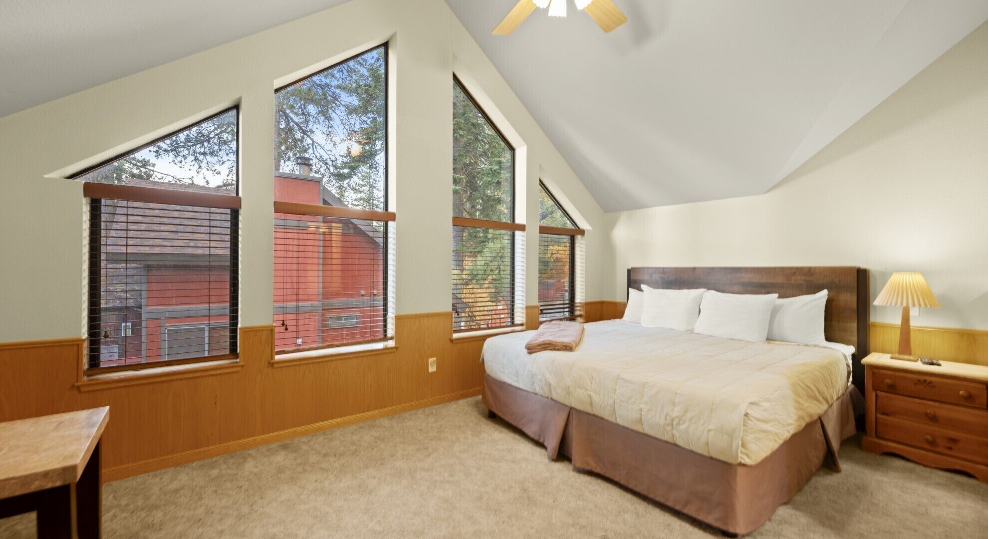 A bedroom with a large bed, a nightstand, a desk, and three large windows.