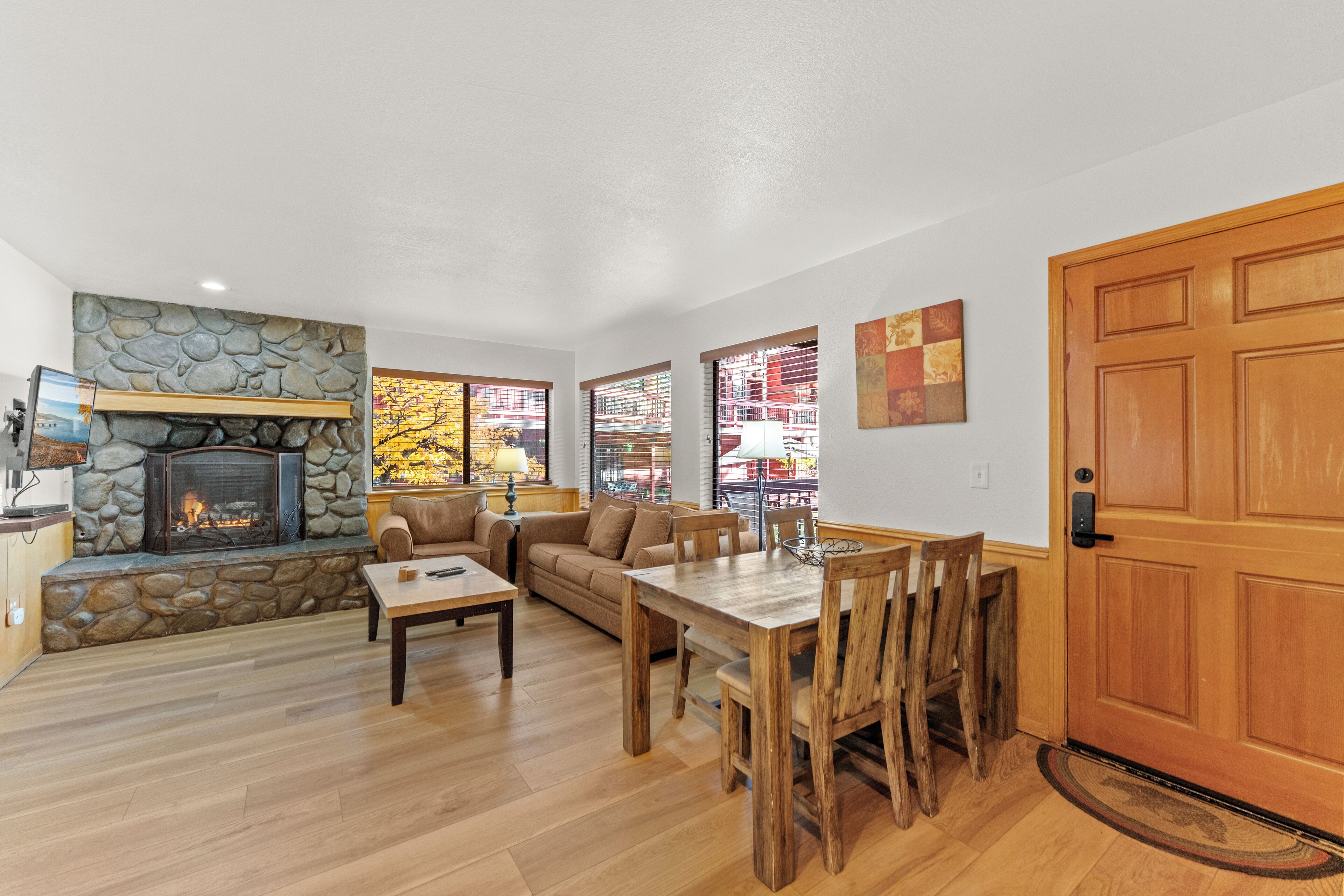 A living room with a stone fireplace, wooden dining table, and a large window with a view of a building.