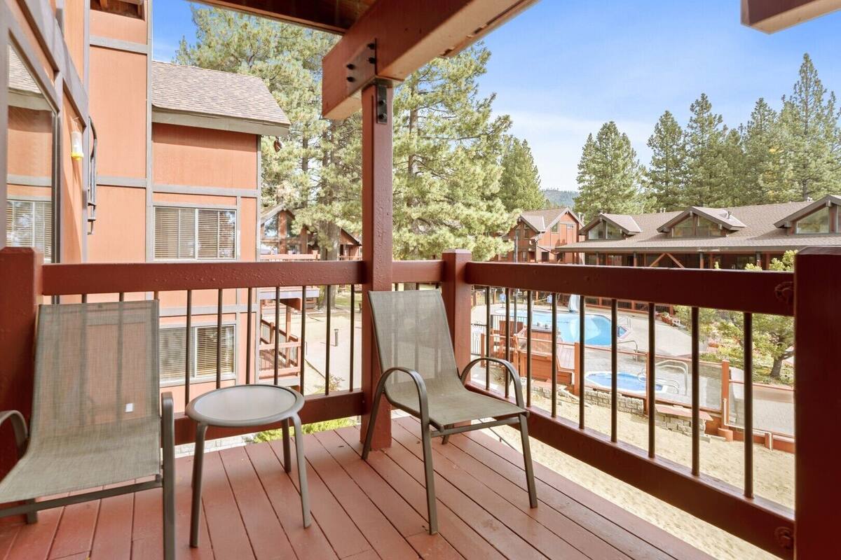 A balcony with two chairs and a table, overlooking a pool and a wooded area.
