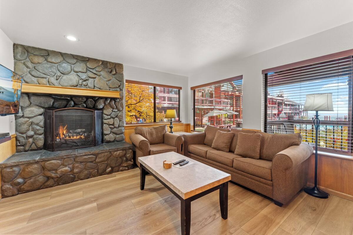 A living room with a stone fireplace, a brown sofa, a coffee table, and large windows with blinds.