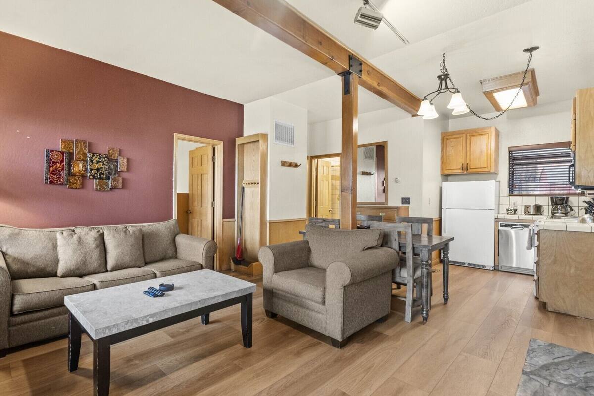 A living room with a sofa, armchair, and coffee table. There is a kitchen area with wooden cabinets and a refrigerator.