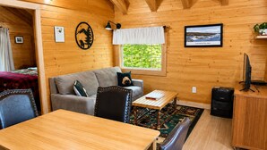 Interior acogedor de una cabaña con una mesa de comedor de madera, un sofá, un televisor pequeño y una ventana con vista a la vegetación.