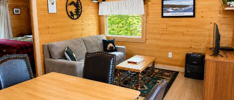 A cozy cabin interior with a wooden dining table, a sofa, a small TV, and a window with a view of greenery.