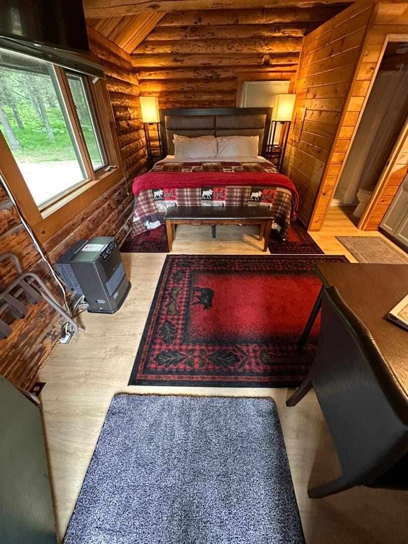 A log cabin room with a bed, a desk, a heater, and a window.