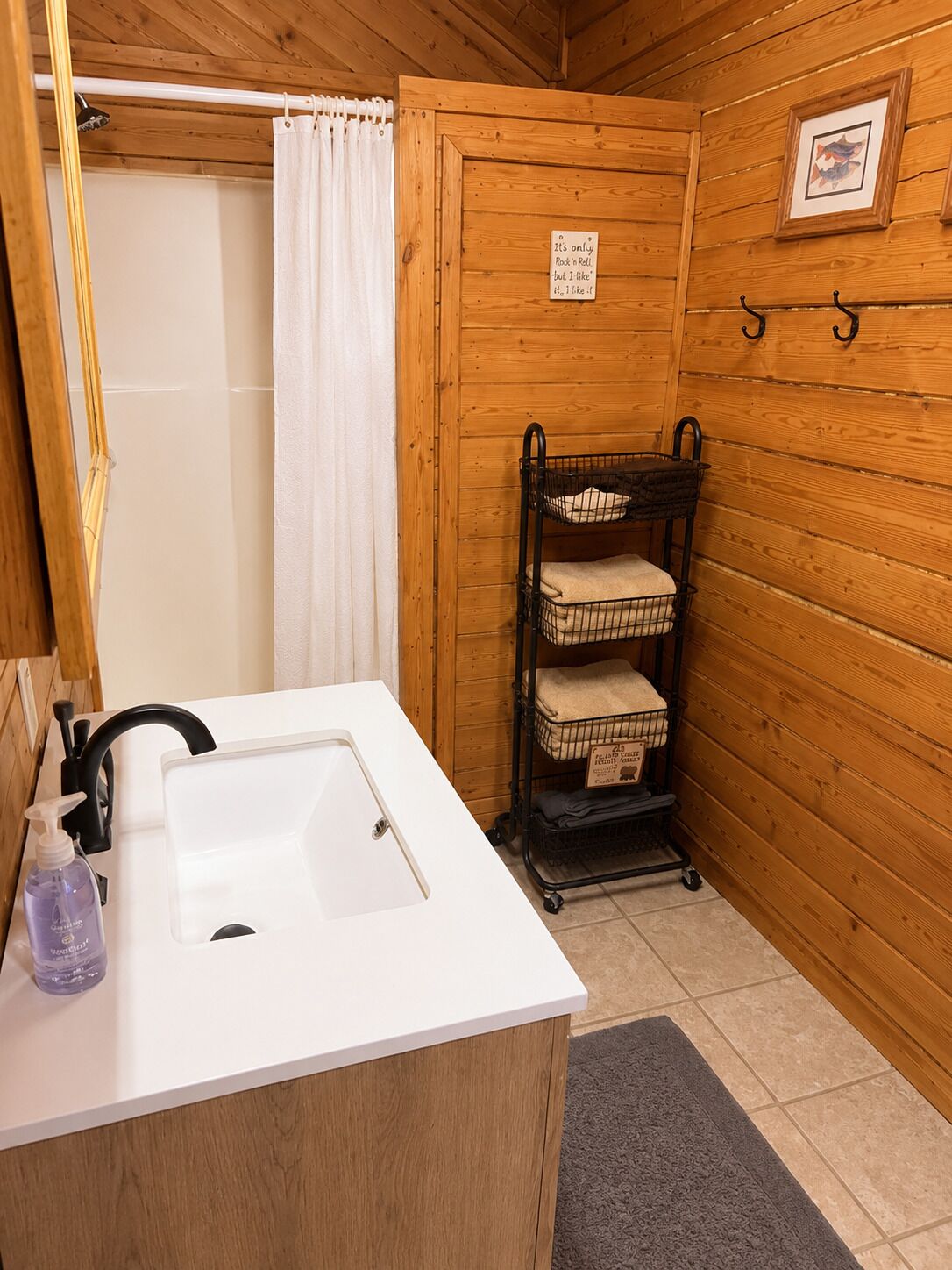 Un bagno in una cabina di legno con un lavandino bianco, una tenda da doccia, un portascopino con asciugamani e un cartello appeso al muro.