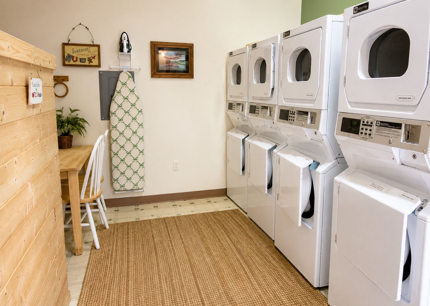 A laundry room with multiple washing machines, a wooden wall, a small table with a plant, and a framed picture on the wall.