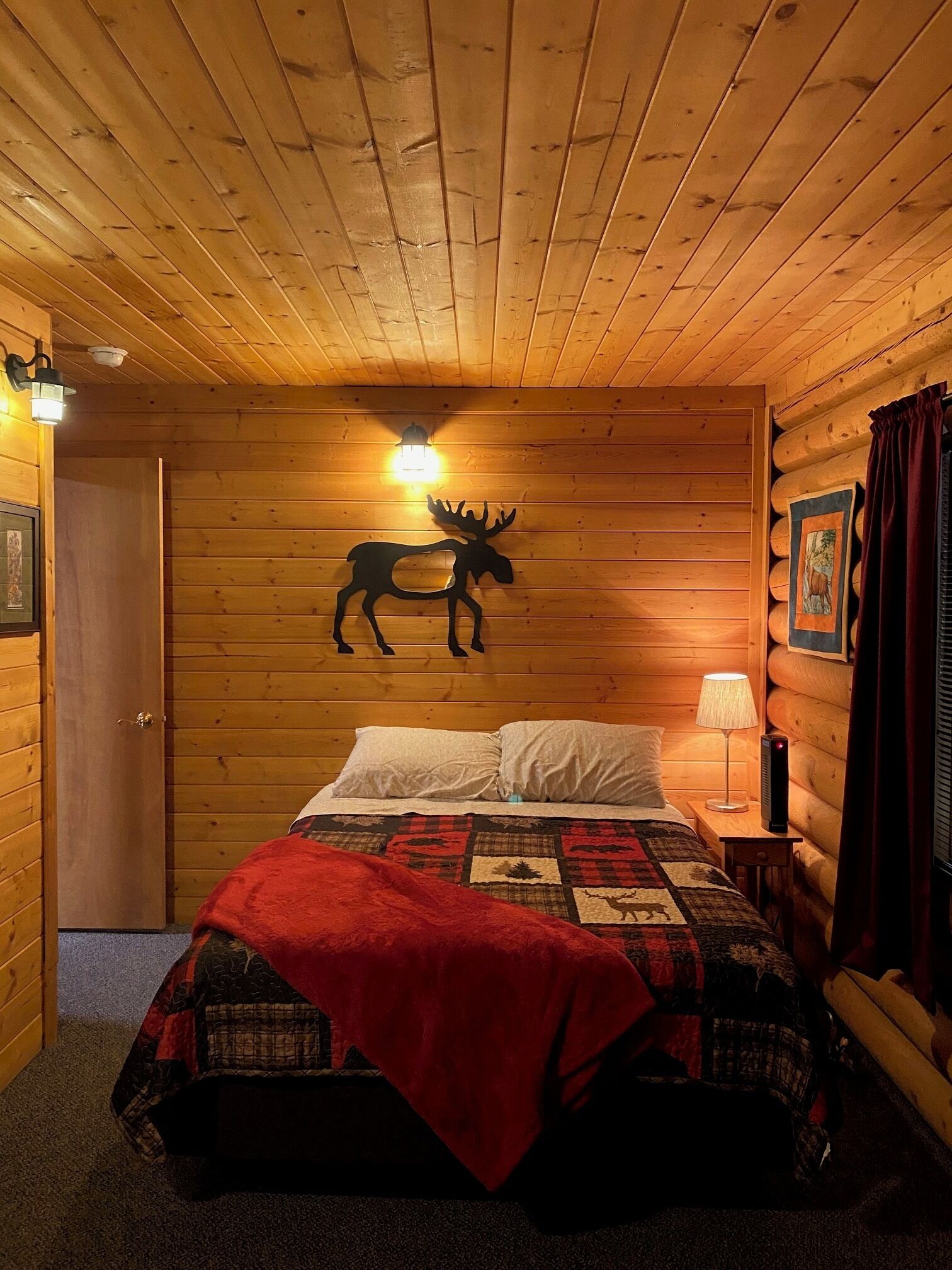 Una habitación tipo cabaña de madera con una cama, un adhesivo de pared con forma de alce, una lámpara y una puerta.