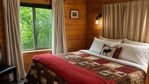 Un dormitorio de cabaña de madera con una cama grande, una ventana con cortinas y un pequeño banco.