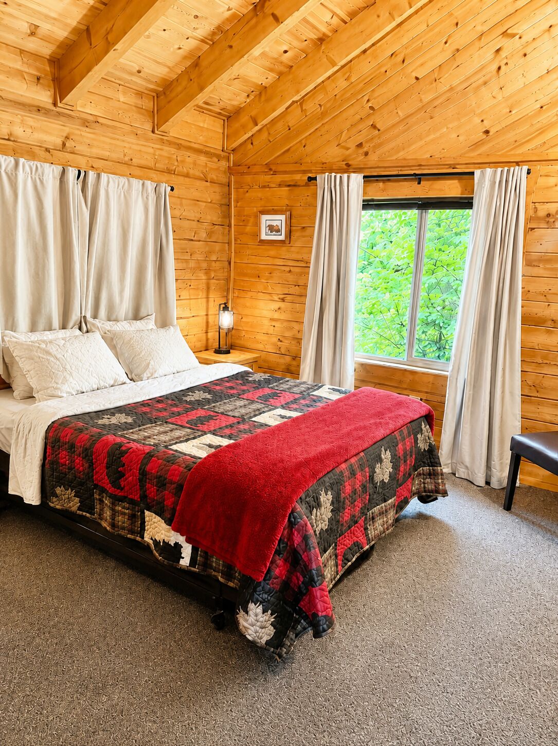 A wooden cabin bedroom with a bed, curtains, a nightstand, a window with a view, and a framed picture on the wall.