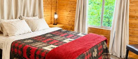 A wooden cabin bedroom with a bed, curtains, a nightstand, a window with a view, and a framed picture on the wall.