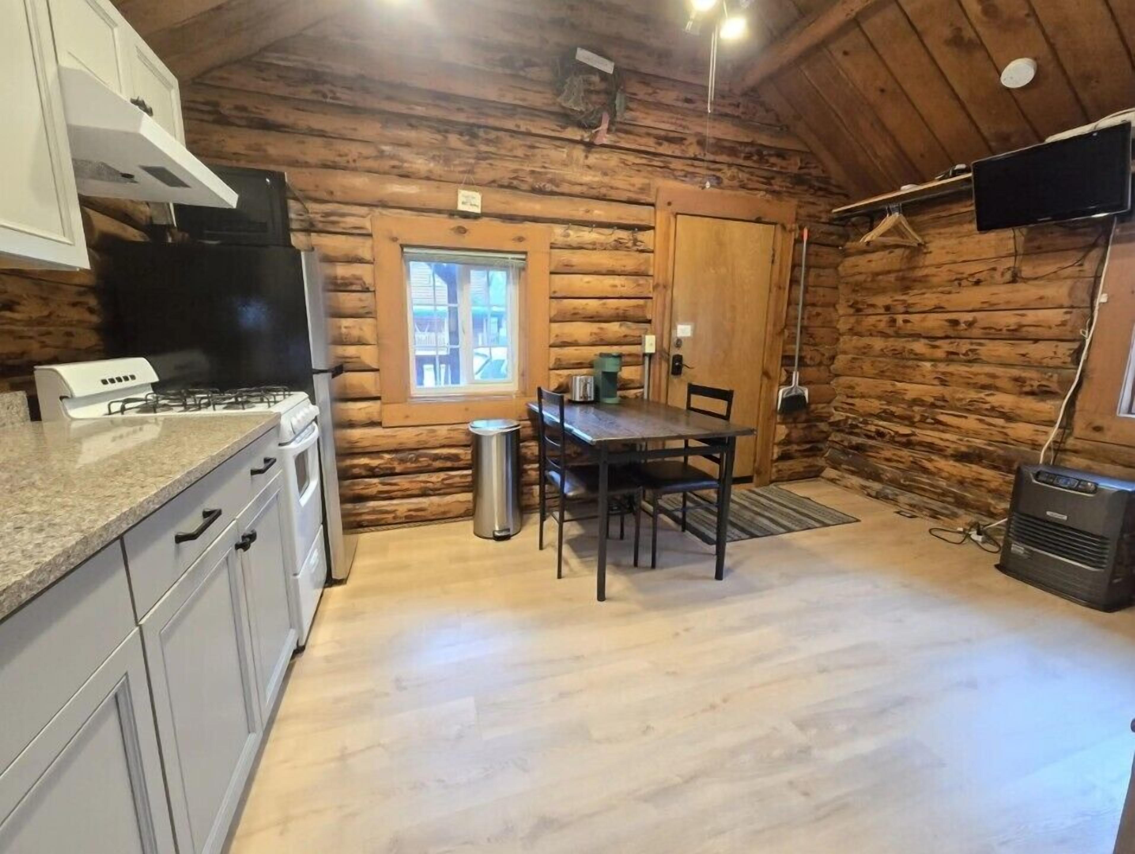 A log cabin interior with a kitchen area, a dining table, and a TV mounted on the wall.