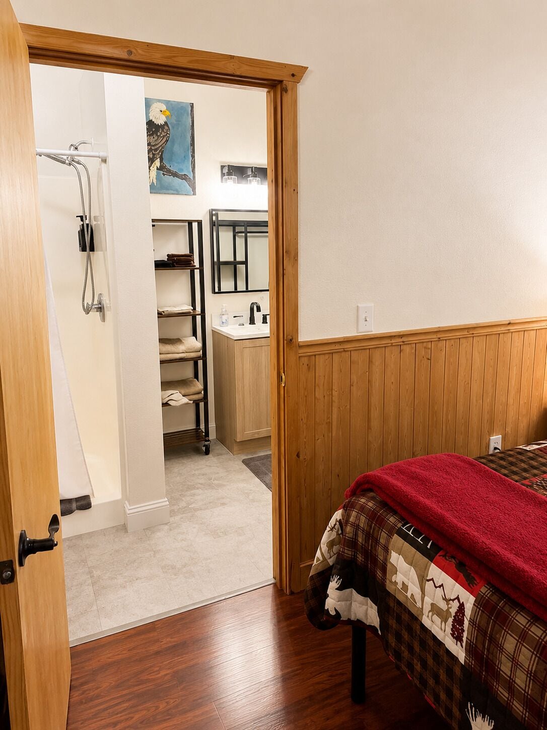A bedroom with a wooden door, a bed with a plaid blanket, a wooden floor, a bathroom with a sink and shelves, and a painting of an eagle.