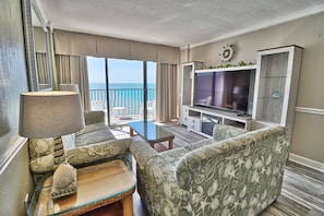 A spacious living room with a large TV, comfortable seating, a balcony with a view of the ocean, and a ship's wheel decoration. - Suites at the Beach (Myrtle Beach)