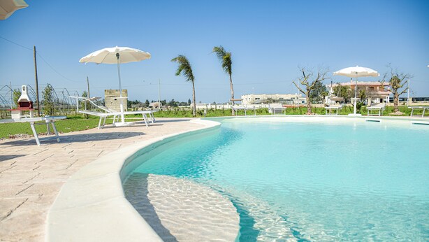 A clear swimming pool with a curved edge, surrounded by lounge chairs and umbrellas. - Il Mondo dei Sogni (Nardò)