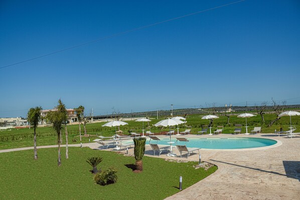 Zona de piscina con sillones de descanso y sombrillas, rodeada de vegetación y un cielo despejado.
