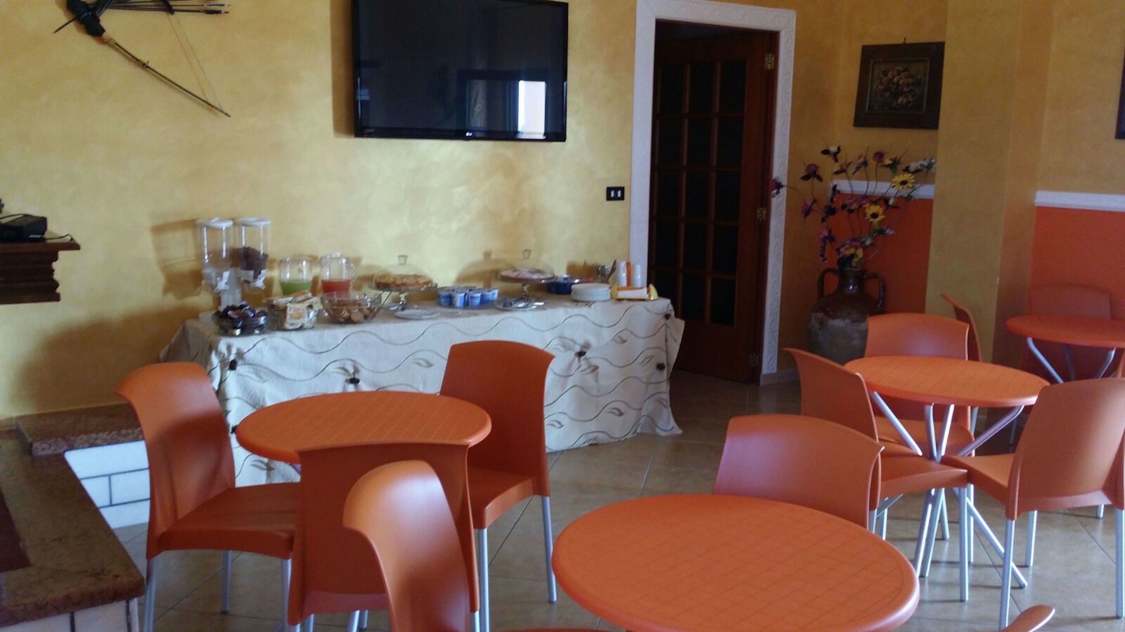 A dining area with orange chairs and tables, a buffet table with various food items, a television mounted on the wall, and a vase with flowers on the wall.