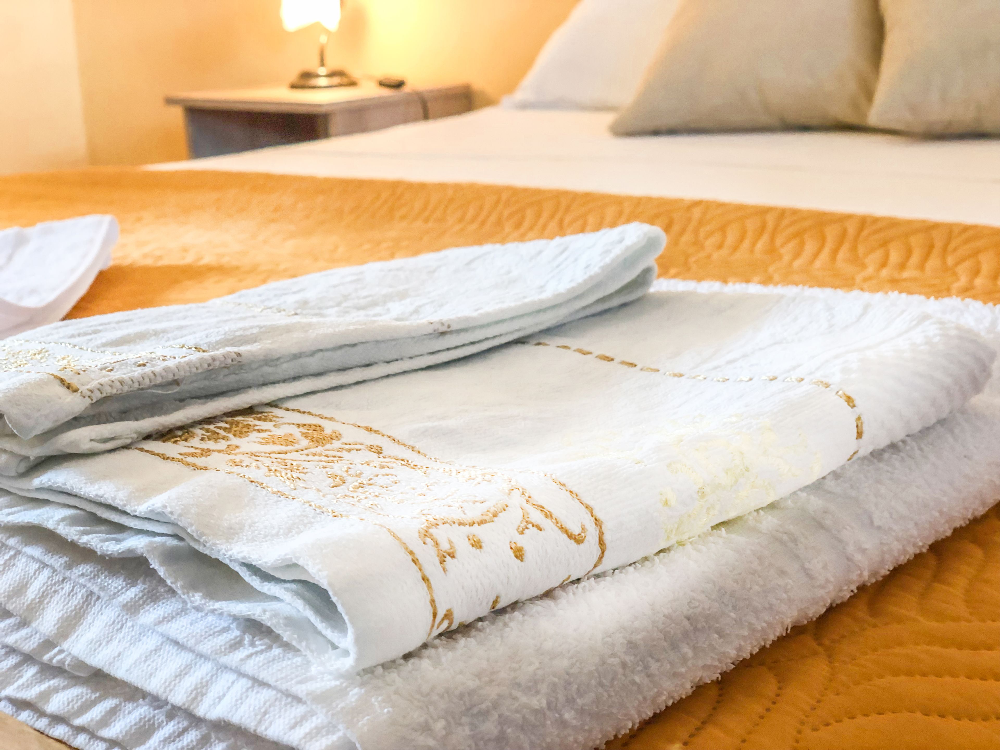 Une serviette blanche, soigneusement pliée et ornée de broderies dorées, est posée sur un lit recouvert d’une couverture orange.