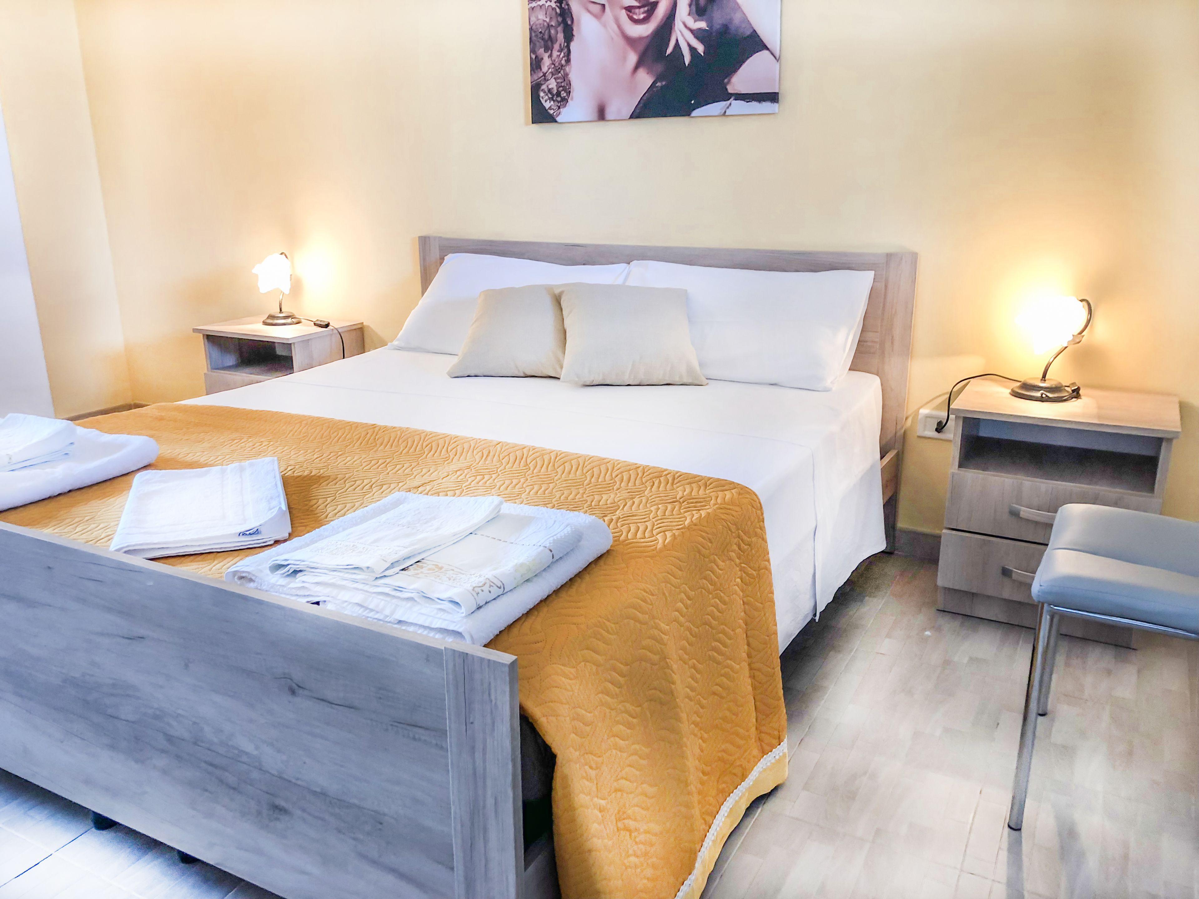 Un lit bien fait, recouvert d’une couverture jaune, avec deux oreillers blancs et des serviettes pliées posées sur le côté. Il y a une table de chevet avec une lampe de chaque côté du lit, et on aperçoit une chaise dans le coin de la pièce.