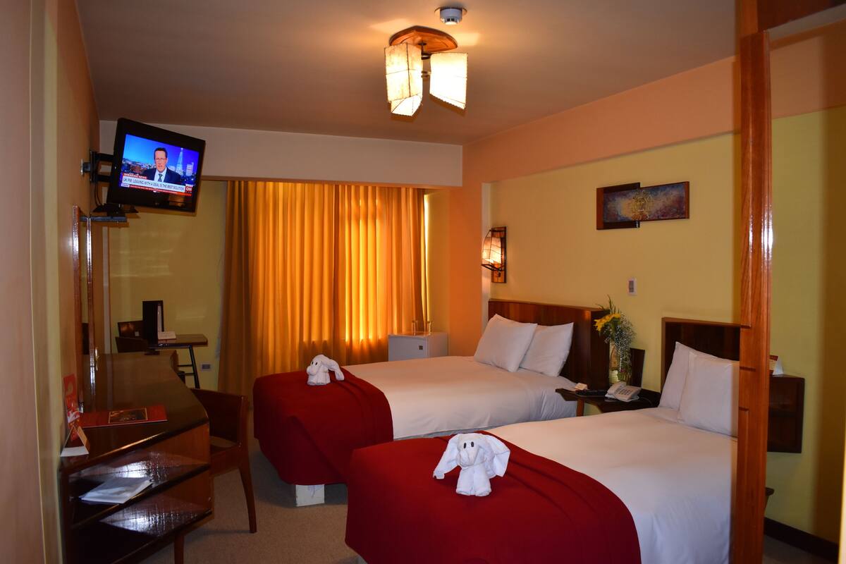 A hotel room with two beds, a TV mounted on the wall, a desk with a chair, and a window with curtains.