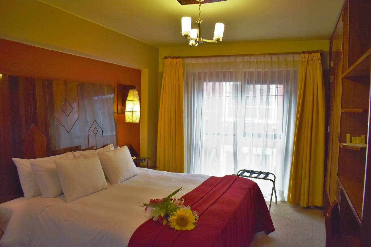 A hotel room with a large bed, a bedside table with a lamp, a window with curtains, and a bookshelf.