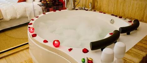 Une chambre d’hôtel avec une grande baignoire blanche entourée de pétales de roses, un lit avec du linge de maison blanc et une table de chevet sur laquelle se trouvent une bouteille de champagne et des verres.
