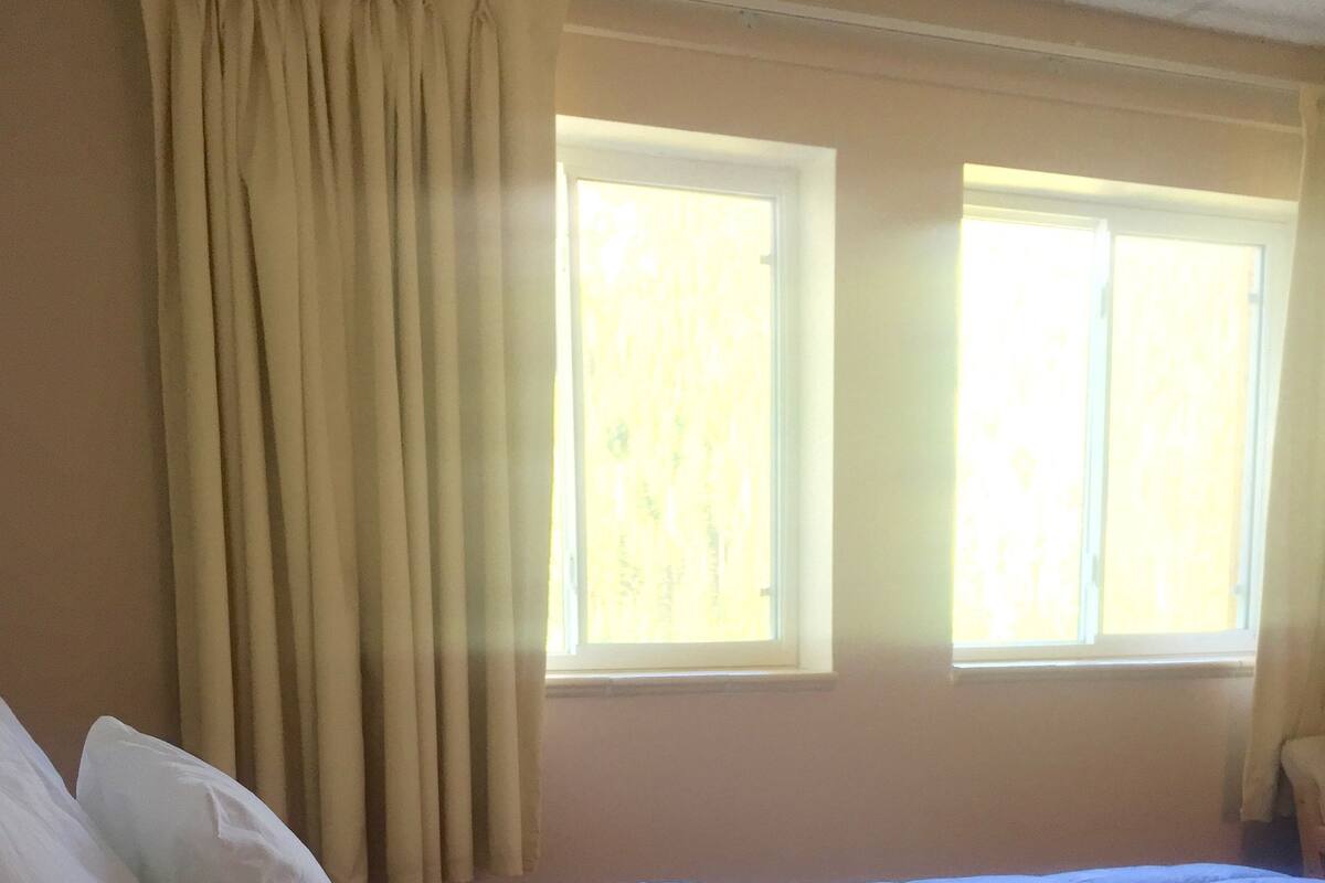A neatly made bed with white linens in a room with two windows and beige curtains.