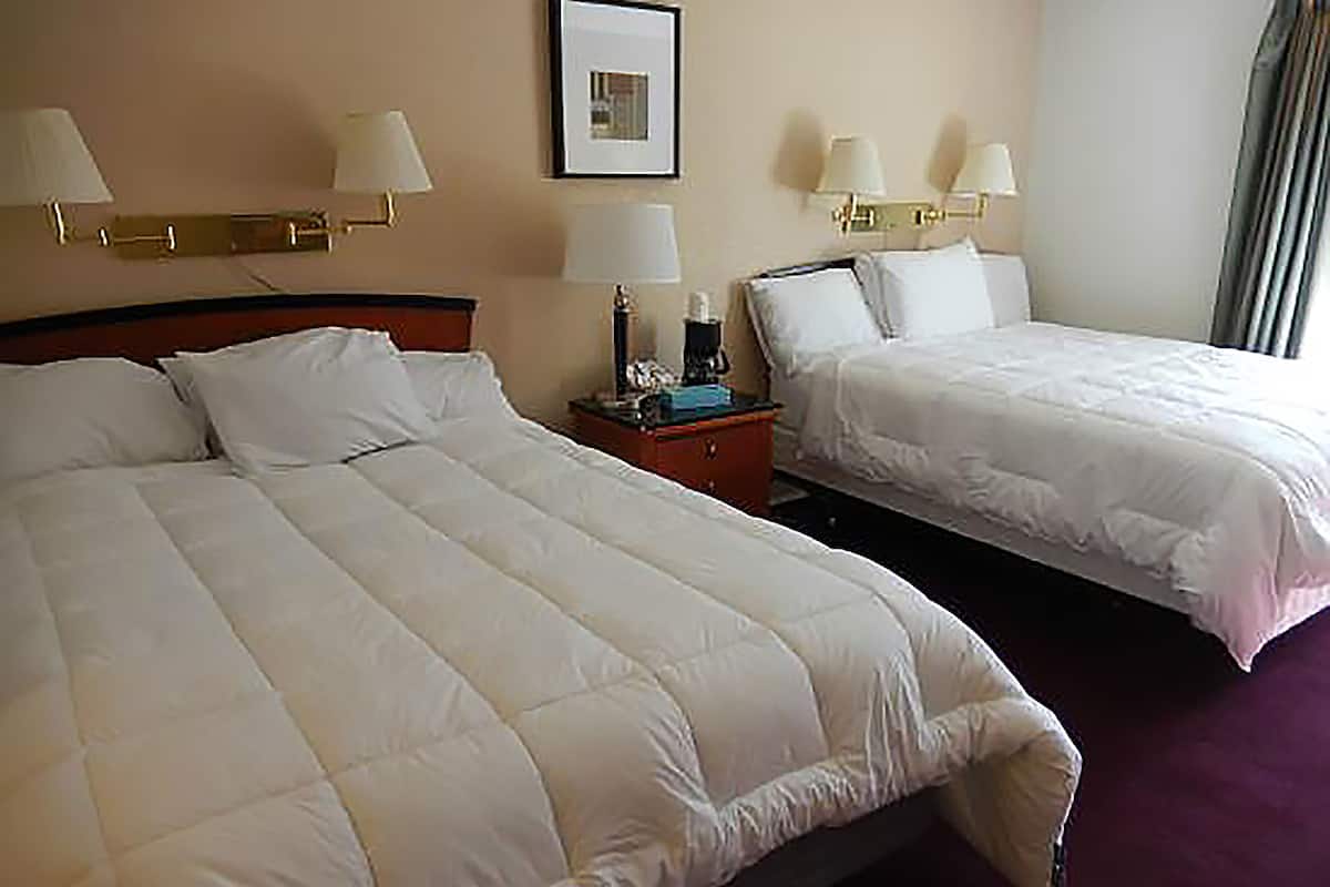 A hotel room with two beds, each with white bedding and pillows. There are two bedside tables with lamps and a framed picture on the wall.