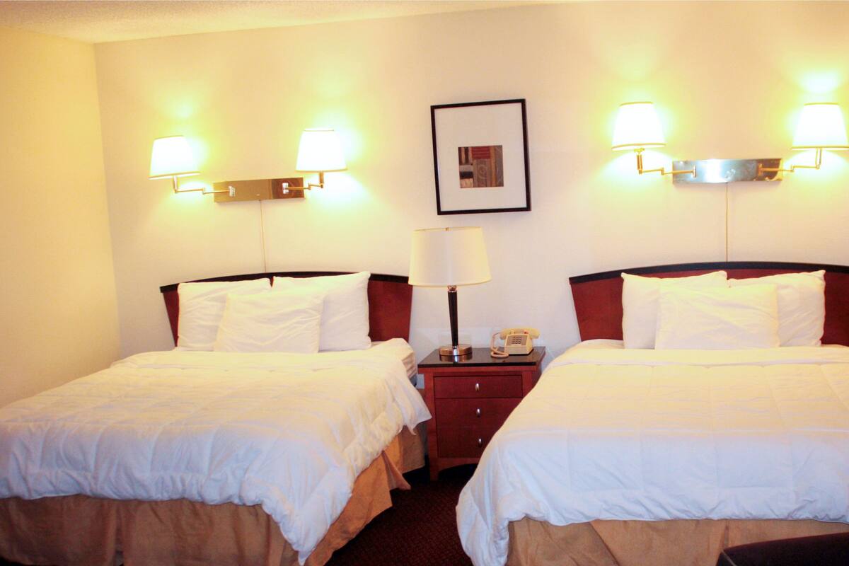 A hotel room with two beds, a nightstand with a lamp, and wall-mounted lights.