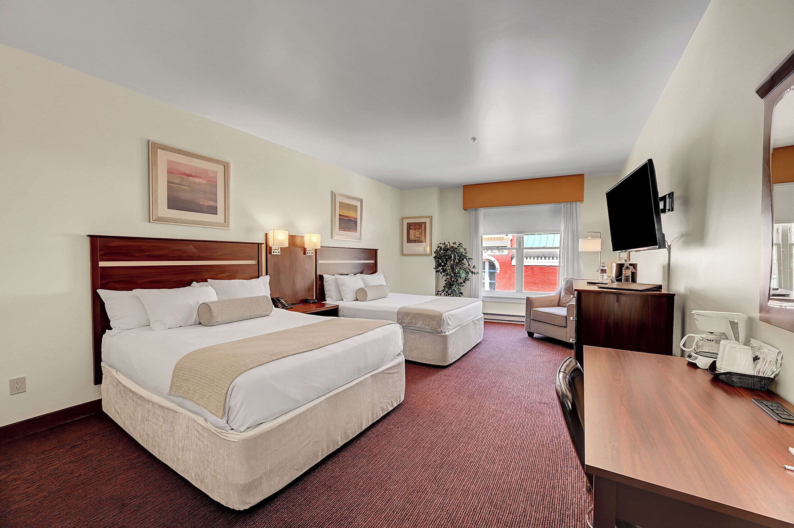 A hotel room with two beds, a desk, a TV, and a window with a view of the outside.