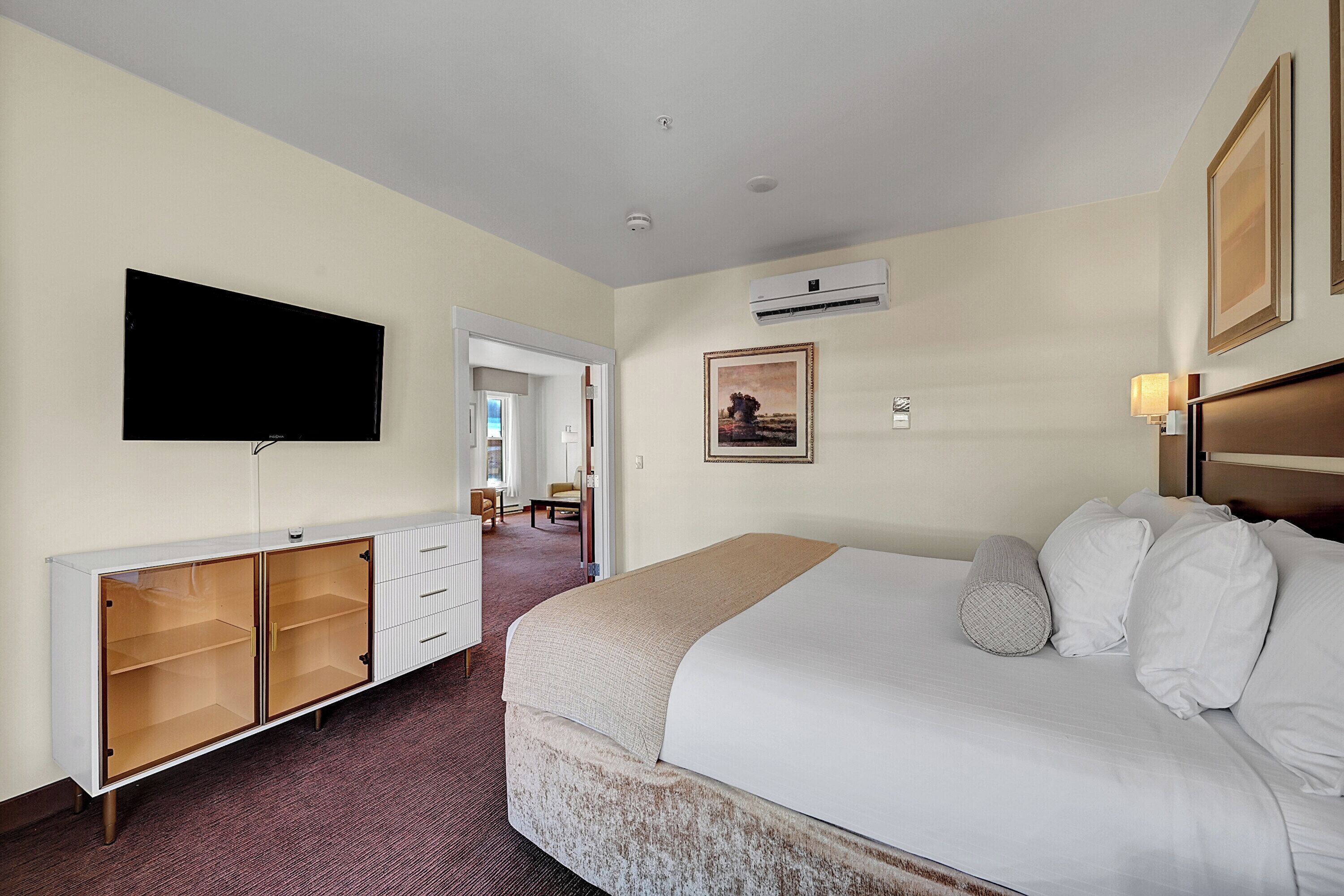 A hotel room with a large bed, a flat-screen TV mounted on the wall, a wardrobe, and an air conditioning unit.