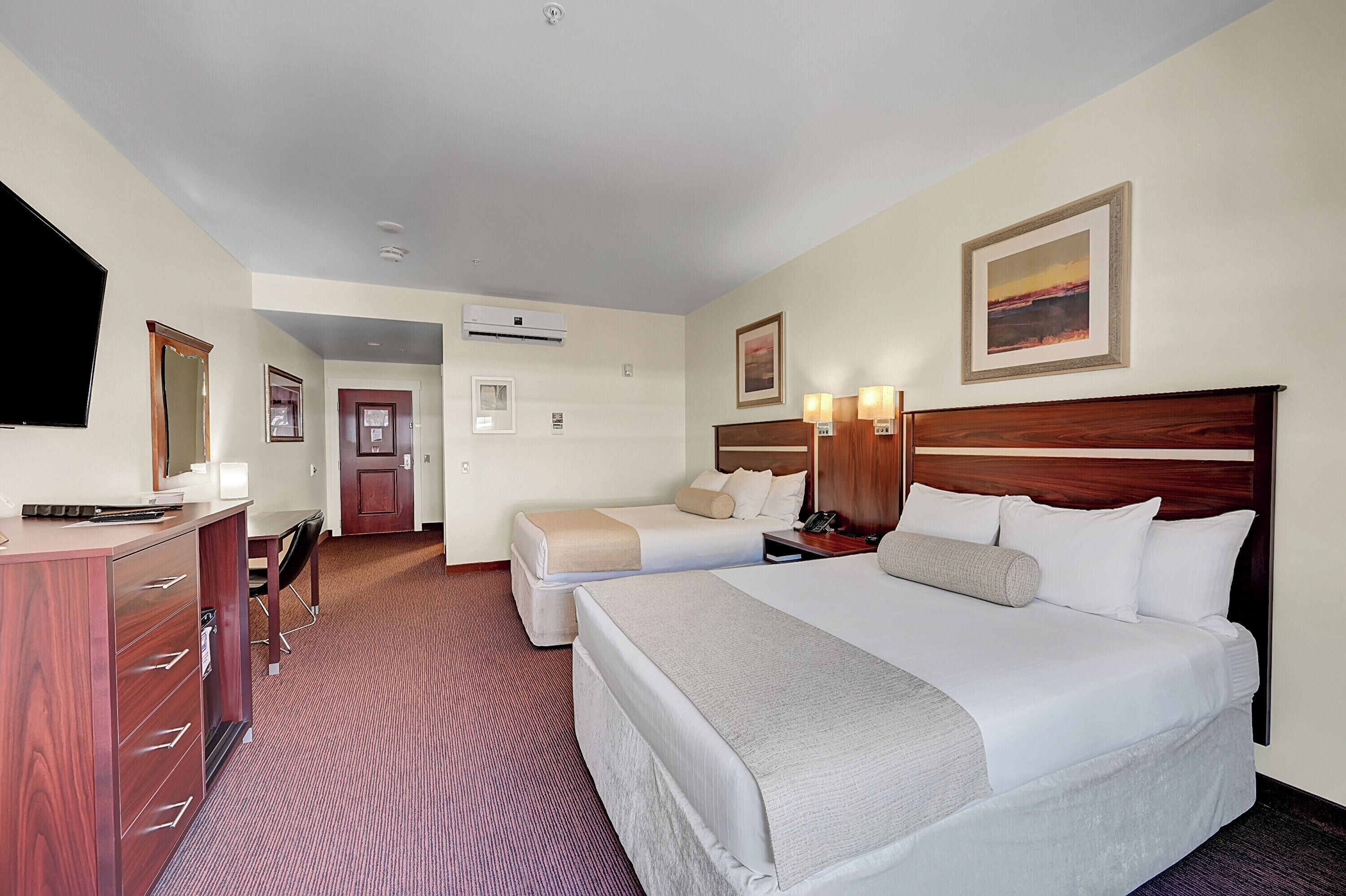 Quarto de hotel com uma cama grande, uma cama menor, uma escrivaninha com gavetas, uma cadeira, uma luminária de mesa, um telefone e um quadro na parede.