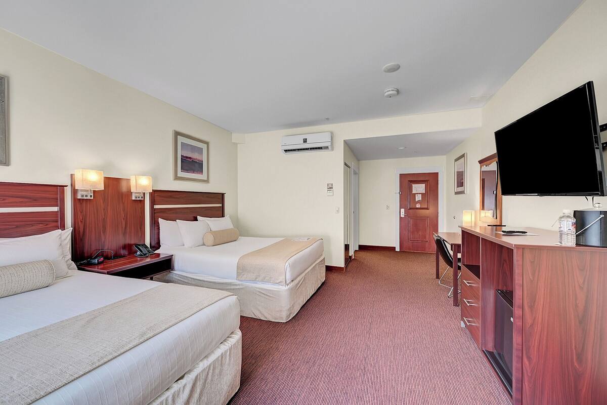 Ein Hotelzimmer mit zwei Betten, einem Schreibtisch, einem Flachbildfernseher, einer Minibar und einer Tür zu einem weiteren Zimmer.